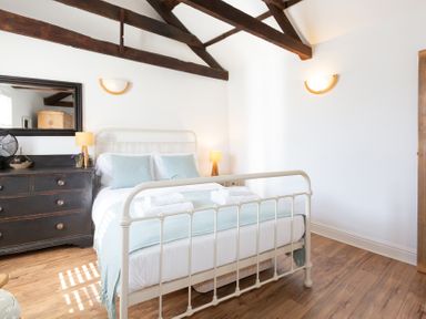 A bedroom with a white metal bed frame wooden beams a black dresser with a mirror and lamps at The Sail Loft in St Ives