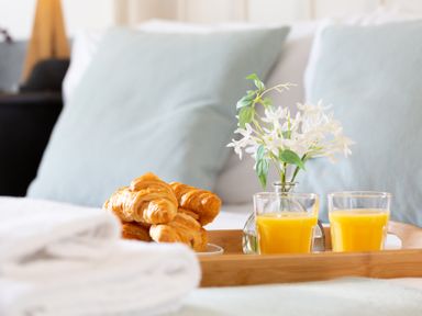 A wooden tray with croissants orange juice and a small vase with white flowers on a bed with pillows at The Sail Loft in St Ives