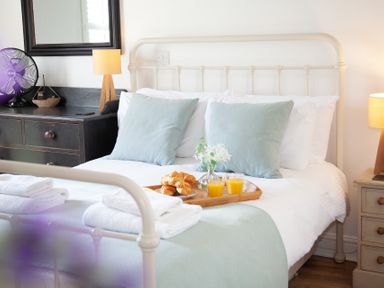 A bedroom with a white metal bed frame blue pillows folded towels and a wooden tray with croissants and glasses of orange juice at The Sail Loft in St Ives
