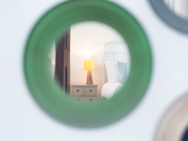 A bedroom with a bedside table and lamp reflected in a green circular mirror at The Sail Loft in St Ives