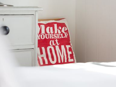 A white bedside drawer next to a pillow with red and white text reading make yourself at home at The Sail Loft in St Ives