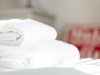 Two folded white towels on a bed with a blurred red cushion in the background at The Sail Loft in St Ives