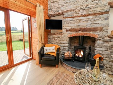 A living room with a stone fireplace wood stove a black armchair and a glass table with wine at Tegfan Barn Pant-y-dwr near Rhayader