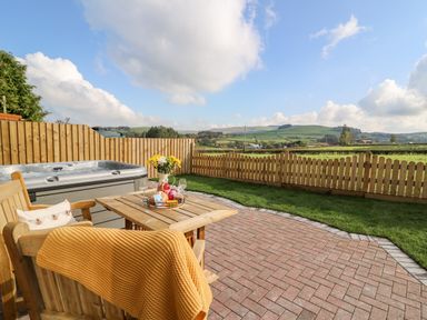 An outdoor patio with wooden chairs a table flowers a hot tub and a fenced grassy area at Tegfan Barn Pant-y-dwr near Rhayader