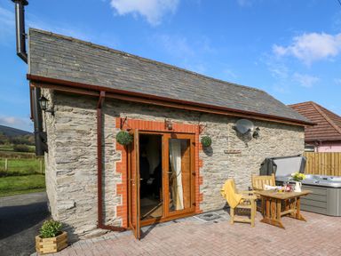 An exterior view of a stone building with open wooden doors a wooden table with chairs and a hot tub at Tegfan Barn Pant-y-dwr near Rhayader