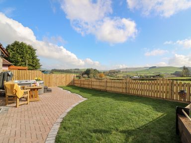 A garden with wooden chairs and table on a paved patio and a fenced lawn with countryside views at Tegfan Barn in Pant-y-dwr near Rhayader