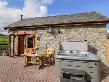 An outdoor patio with a wooden table and chairs and a hot tub outside a stone building at Tegfan Barn Pant-y-dwr near Rhayader