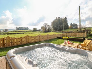 An outdoor hot tub with bubbling water next to a wooden table with flowers and drinks and wooden chairs in a fenced garden with fields and trees beyond at Tegfan Barn in Pant-y-dwr near Rhayader