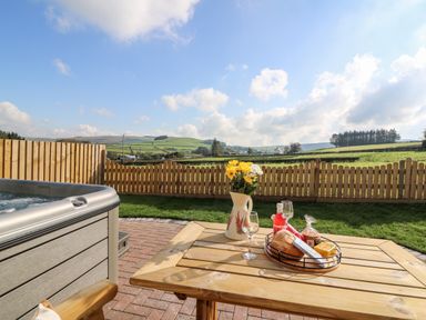 A garden area with wooden table holding flowers and food next to a hot tub and wooden fence at Tegfan Barn in Pant-y-dwr near Rhayader