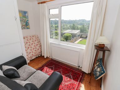 A small room with a gray and black sofa a red patterned rug a wooden floor a window with white curtains and a lamp on a wooden stand at Hawthorn House in Pembroke