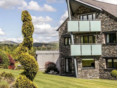 A stone building with glass balconies and a spiral trimmed bush on a grass lawn at The Golf Apartment in Kendal