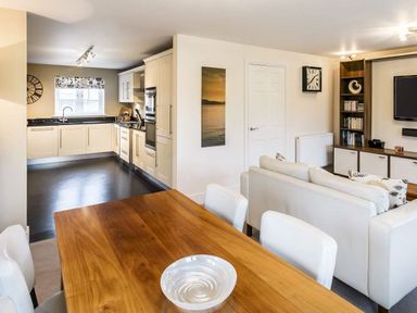 An open plan living space with a wooden dining table and white chairs a white sofa a built in shelving unit a wall clock and a kitchen with cream cabinets at The Golf Apartment in Kendal