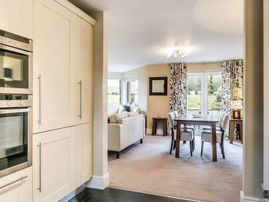 A dining area with wooden table and four chairs near glass doors with floral curtains and a living room with white sofas visible at The Golf Apartment in Kendal
