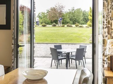 A dining area with a bowl on the table looking out through open double doors onto a patio with a table and chairs and a grassy area beyond with people playing golf at The Golf Apartment in Kendal