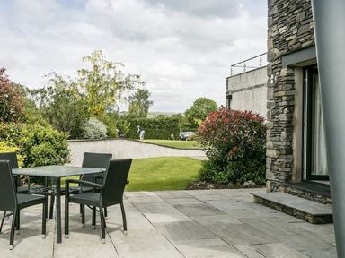 An outdoor patio with a table and four chairs next to a stone building and a garden with trees and shrubs at The Golf Apartment in Kendal