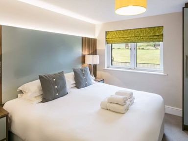 A bedroom with a double bed with grey pillows and folded white towels on top a bedside lamp window with a green blind at The Golf Apartment in Kendal