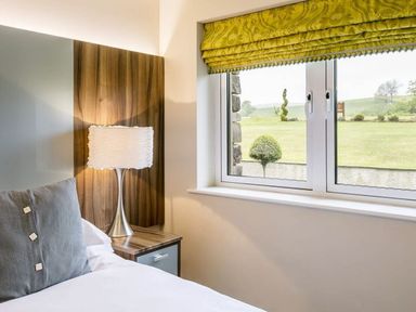 A bedroom corner with a bed pillow a bedside table with a lamp and a window with green Roman blinds at The Golf Apartment in Kendal