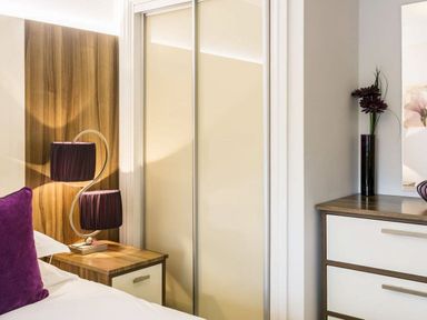 A bedroom corner with a wooden nightstand holding a lamp with two purple shades and a purple pillow on the bed at The Golf Apartment in Kendal