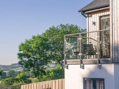 A balcony with two chairs and a small table on a wooden house overlooking trees and distant hills at Swallows Dip in Honiton