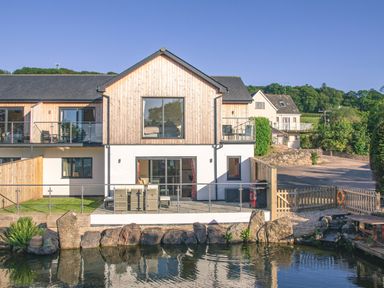 A two story building with wooden and white exterior walls overlooking a pond with rocks and outdoor furniture on a patio at Swallows Dip in Honiton