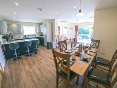 A dining table set for six next to an open kitchen with bar stools and a seating area with sliding glass doors at Swallows Dip in Honiton
