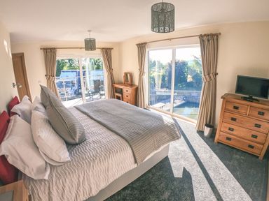 A bedroom with a bed pillows wooden dressers a television and large windows with curtains at Swallows Dip in Honiton