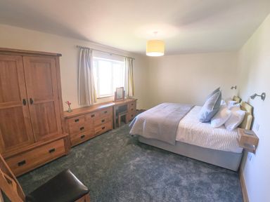 A bedroom with a bed, wooden wardrobe, dresser with mirror, curtains, and a chair at Swallows Dip in Honiton