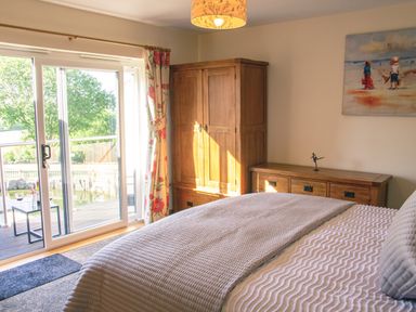A bedroom with a bed a wooden wardrobe a dresser and a sliding glass door showing a deck and trees outside at Swallows Dip in Honiton