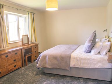 A bedroom with a bed and pillows a wooden dresser with mirror and chair and curtains on a window at Swallows Dip in Honiton