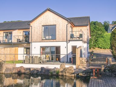 An exterior view of a two-story house with wooden paneling and a glass balcony by a pond at Swallows Dip in Honiton