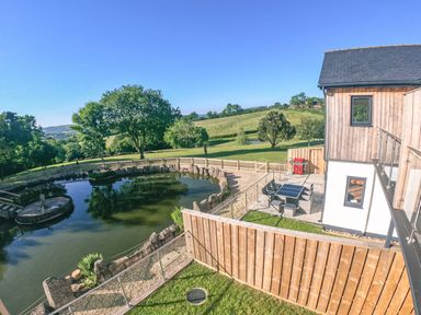 An outdoor patio with a table and chairs next to a house and a pond with a wooden fence and trees at Swallows Dip in Honiton