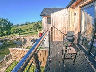 A balcony with a chair and a small table holding a wine bottle and glasses overlooking a fenced pond and green fields at Swallows Dip in Honiton
