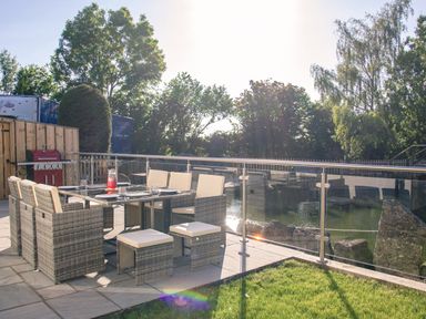 An outdoor patio with a dining table and chairs next to a glass railing overlooking a pond at Swallows Dip in Honiton