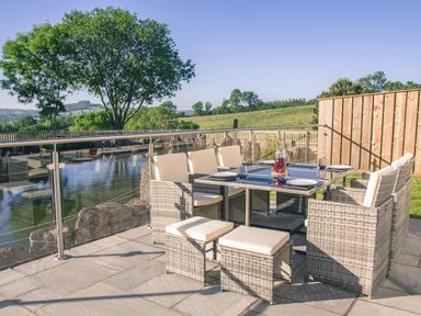An outdoor patio dining area with seven wicker chairs and a glass table set with plates and glasses overlooking a pond and countryside at Swallows Dip in Honiton