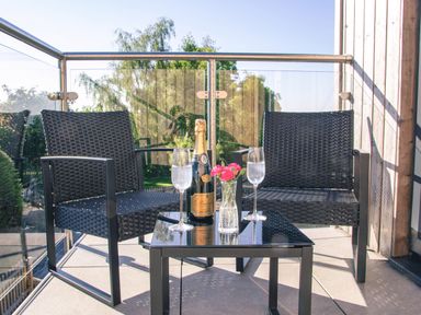 A balcony with two black woven chairs and a black table holding a champagne bottle two glasses and a vase with pink flowers at Swallows Dip in Honiton