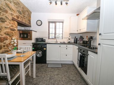 A kitchen with white cabinets a black washing machine a microwave a wooden dining table with chairs and stone wall at Trem Y Wyddfa in Gaerwen
