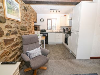 A kitchen with white cabinets and black countertops next to a dining table and a gray armchair with a pillow at Trem Y Wyddfa in Gaerwen