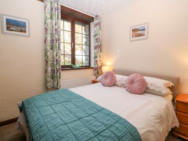 A bedroom with a bed covered with white and teal bedding and pink round pillows by a window with floral curtains at Trem Y Wyddfa in Gaerwen