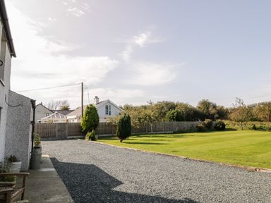 A gravel driveway next to a lawn with trees and houses behind a wooden fence at Penllyn in Newborough