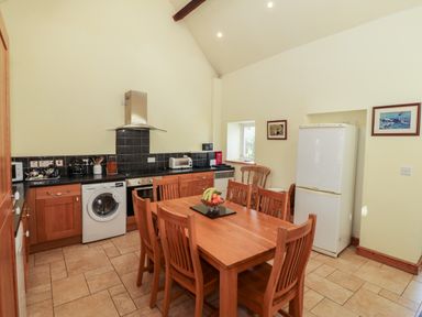 A kitchen with wooden cabinets a refrigerator a washing machine a stove a dining table with six chairs and a fruit bowl at Penllyn in Newborough