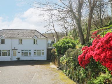 A white house with multiple windows next to a stone garden wall with red flowers and trees at Longships in Porthcurno