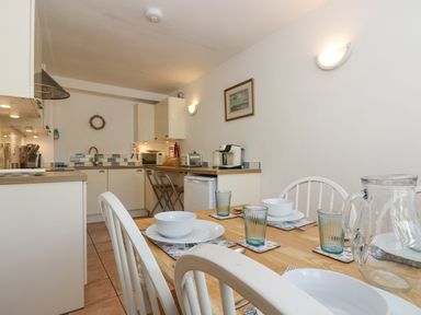 A kitchen and dining area with a wooden table set with bowls and glasses and a kitchen with white cabinets at Longships in Porthcurno