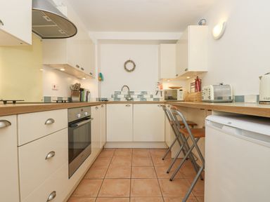 A kitchen with cream cabinets a built-in oven two folding chairs and small appliances at Longships in Porthcurno