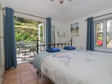A bedroom with a metal bed frame blue curtains and a door opening to a patio with chairs at Longships in Porthcurno