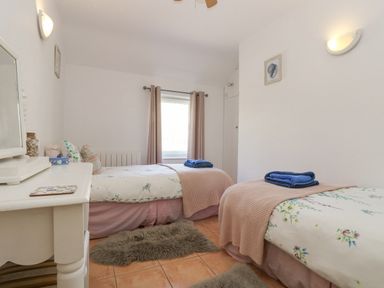 A bedroom with two single beds a window with curtains a white dresser and fur rugs on the floor at Longships in Porthcurno