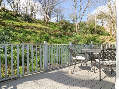 An outdoor deck with metal chairs and a table overlooking a garden with trees and shrubs at Longships in Porthcurno