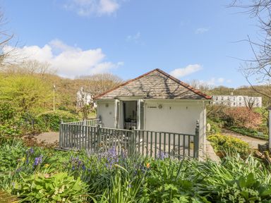 A single story house with a fenced patio and open doors surrounded by plants and trees at Longships in Porthcurno