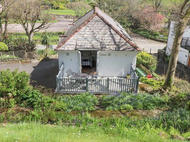 A small house with a fenced patio and outdoor table and chairs surrounded by plants and trees at Longships in Porthcurno