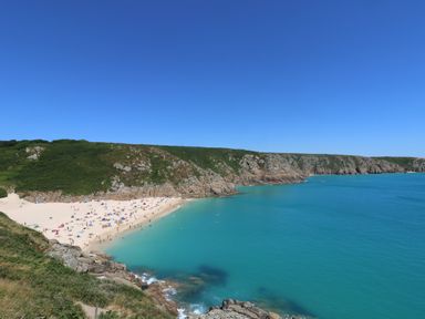 A sandy beach with people and umbrellas next to turquoise water and rocky cliffs covered with vegetation at Longships in Porthcurno