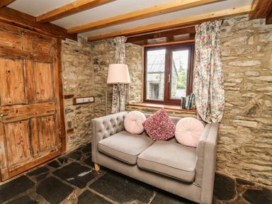 A room with stone walls wooden door window with floral curtains a gray sofa with cushions and a floor lamp at Pant Y Rhedyn Abbeycwmhir near Rhayader